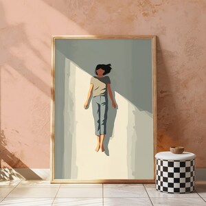 May include: A framed wall art print featuring a stylized illustration of a person with dark hair, wearing a light brown t-shirt and blue pants, floating in a vertical beam of light against a muted sage green background with sharp shadows.