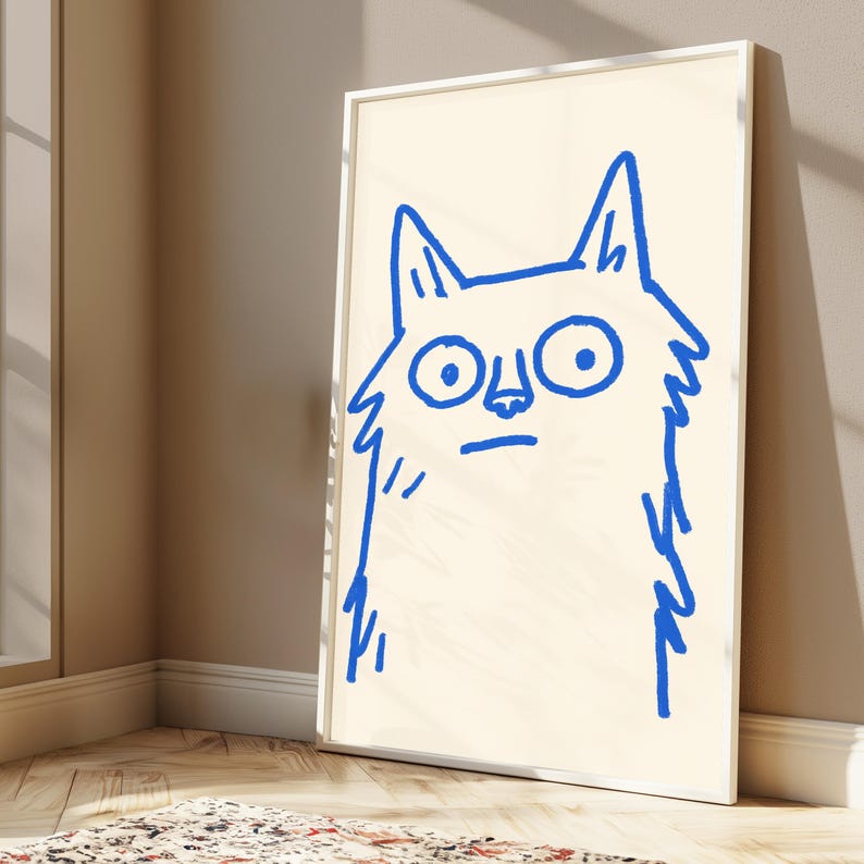 May include: A framed wall art print featuring a simple, hand-drawn illustration of a cat in vibrant blue ink on a cream-colored background. The cat has large, wide eyes, pointed ears, and a straight line for a mouth, rendered in a loose, sketchy style.