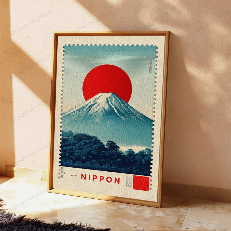 K&ouml;nnte beinhalten: Ein gerahmter Kunstdruck im Design einer Briefmarke mit einer gro&szlig;en roten Sonne &uuml;ber einem schneebedeckten Berggipfel, dunkelblauen B&auml;umen am Fu&szlig; des Berges und dem in Rot gedruckten Schriftzug NIPPON mit einem kleinen roten Quadrat daneben.