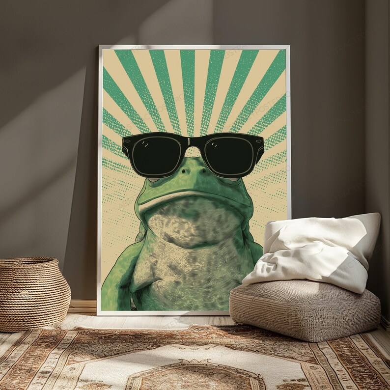 May include: A rectangular wall art print featuring a green frog wearing large black sunglasses. The background consists of a light beige and green sunburst pattern with a dotted texture, creating a retro graphic design aesthetic.