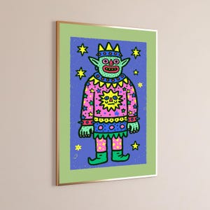 May include: A framed art print featuring a light green creature with pointed ears and a crown, wearing a pink sweater with a yellow sun graphic and star patterns. The figure stands against a blue background filled with yellow stars, all enclosed in a light green border and a thin gold frame.