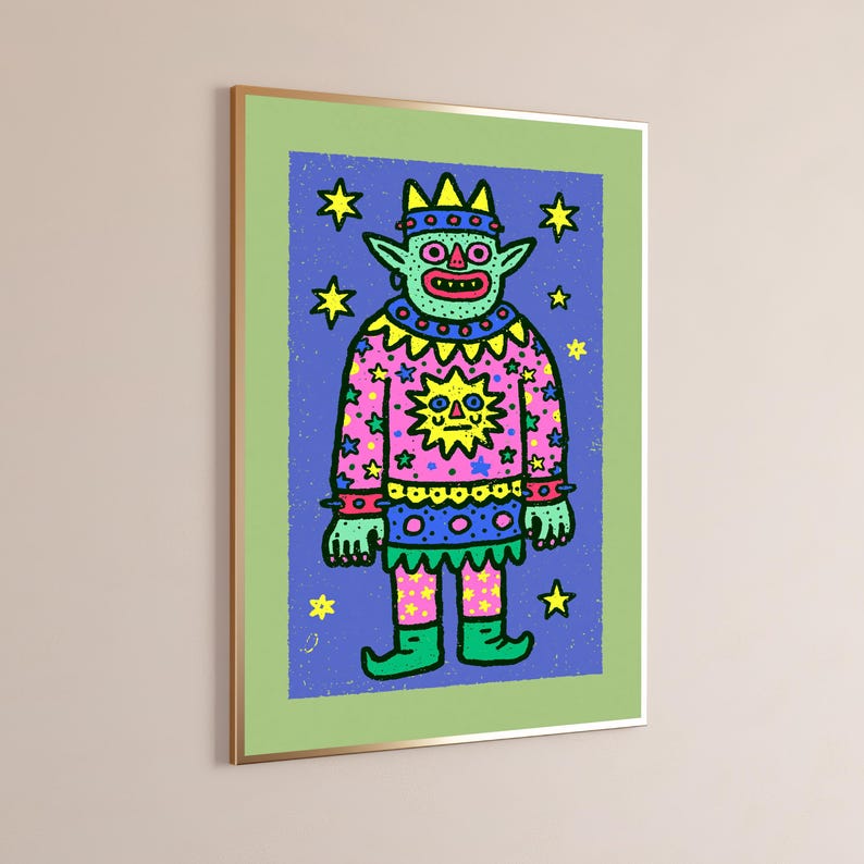 May include: A framed art print featuring a light green creature with pointed ears and a crown, wearing a pink sweater with a yellow sun graphic and star patterns. The figure stands against a blue background filled with yellow stars, all enclosed in a light green border and a thin gold frame.