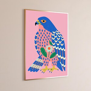May include: A framed wall art print featuring a stylized falcon illustration against a solid pink background. The bird has a blue head and wings with yellow and white accents, a pink chest with blue spots, and a central white flower graphic.