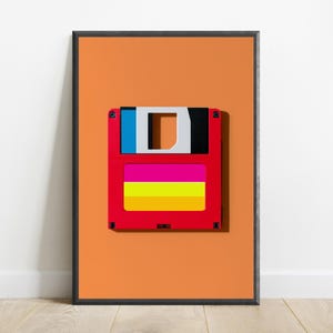 May include: A framed wall art print featuring a centred graphic of a red floppy disk against a solid orange background. The disk has a blue and black top section, with a label area displaying horizontal stripes in bright pink, yellow, and orange.