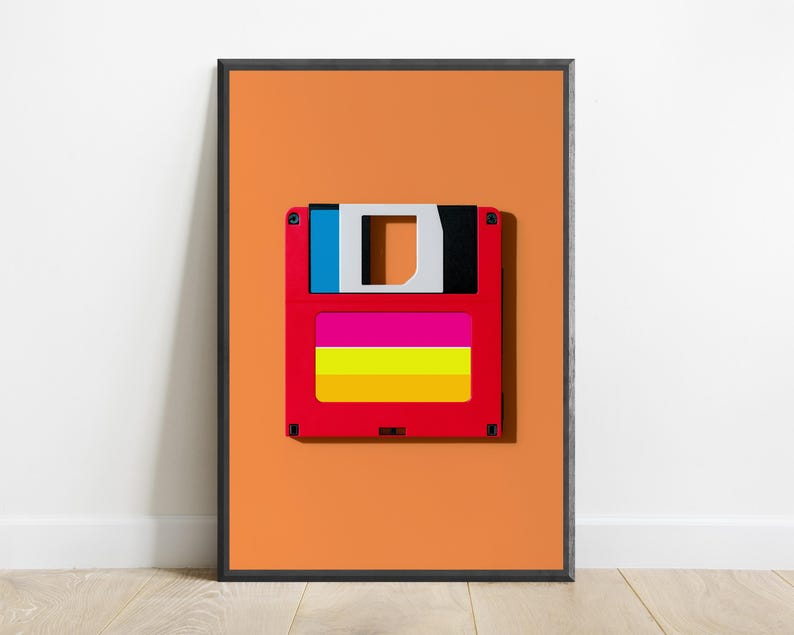 May include: A framed wall art print featuring a centred graphic of a red floppy disk against a solid orange background. The disk has a blue and black top section, with a label area displaying horizontal stripes in bright pink, yellow, and orange.