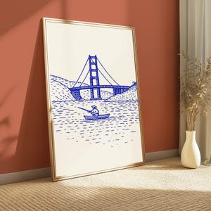 May include: A framed vertical wall art print featuring a blue line drawing on a cream background. The illustration depicts a person in a small boat fishing on the water with the Golden Gate Bridge visible in the background between two hills.