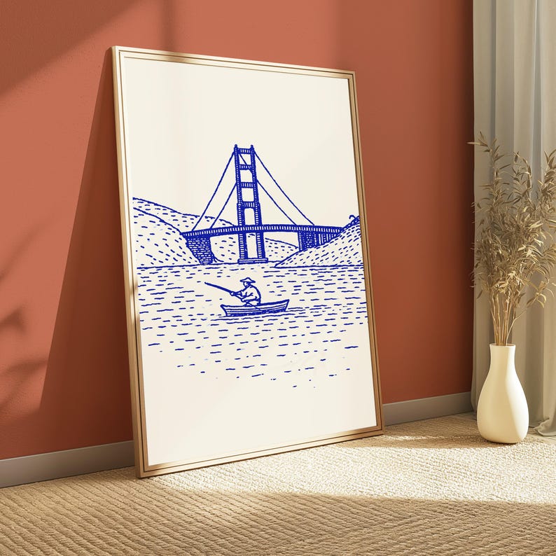 May include: A framed vertical wall art print featuring a blue line drawing on a cream background. The illustration depicts a person in a small boat fishing on the water with the Golden Gate Bridge visible in the background between two hills.