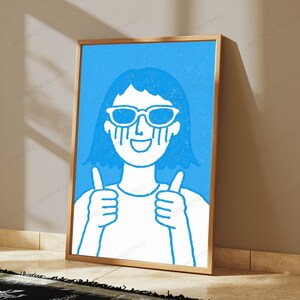 May include: A framed wall art print featuring a white line drawing of a person with short hair wearing sunglasses and giving a double thumbs up gesture, set against a solid bright blue background with a textured, grainy artistic finish.