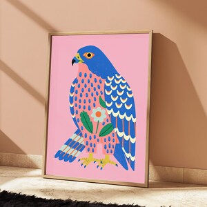 May include: A framed art print featuring a stylized blue falcon on a solid pink background. The bird has a patterned chest with pink and blue spots, a single white flower with an orange center, and yellow talons. The wings display blue feathers.