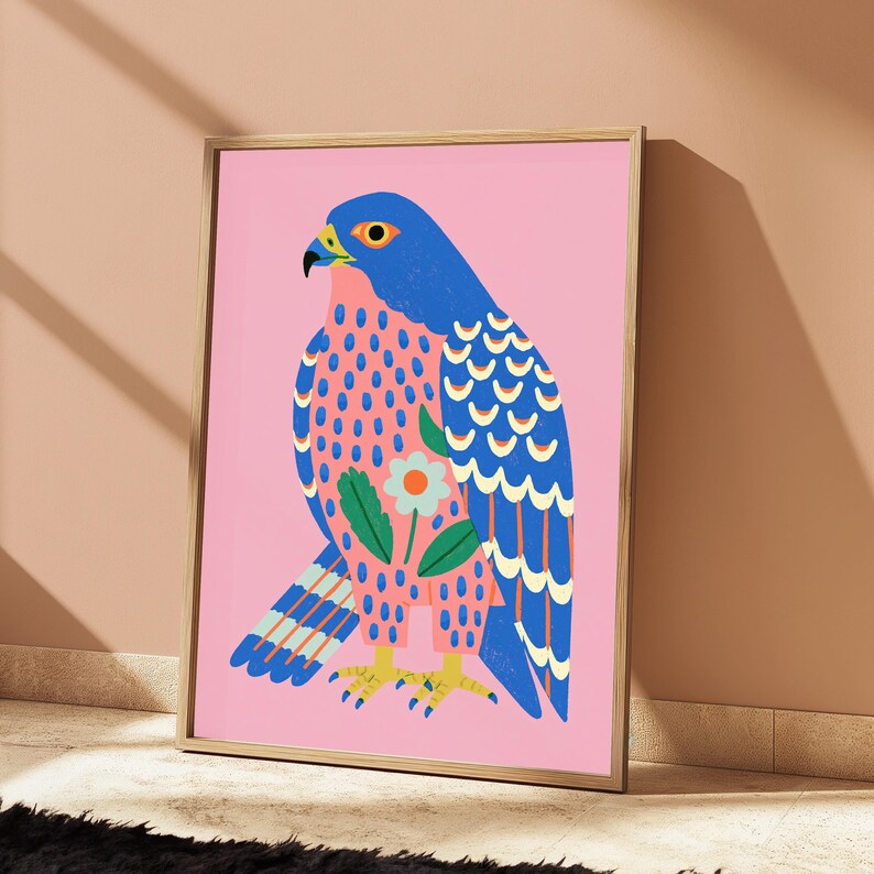 May include: A framed art print featuring a stylized blue falcon on a solid pink background. The bird has a patterned chest with pink and blue spots, a single white flower with an orange center, and yellow talons. The wings display blue feathers.