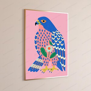 May include: A framed wall art print featuring a stylized blue falcon against a solid pink background. The bird has a yellow beak, orange eye, and a patterned chest with a central white flower and green leaves. The wings display blue and white feathers.