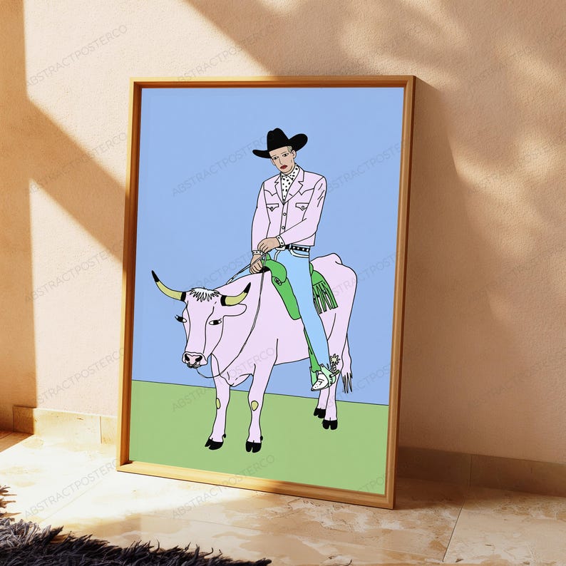 May include: A framed art print featuring a line-art illustration of a person wearing a black cowboy hat, a light pink jacket, and blue jeans, riding a light pink bull with yellow horns. The background consists of a light blue sky and green grass.
