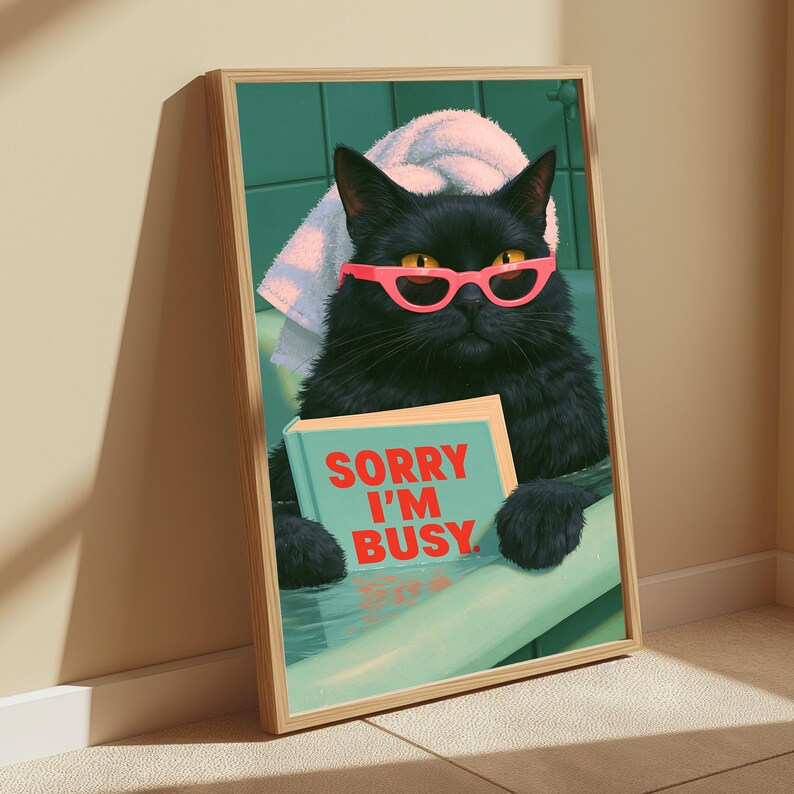 May include: A framed wall art print featuring a black cat with yellow eyes wearing bright pink glasses and a white towel on its head while sitting in a bathtub. The cat holds a light blue book with the red text SORRY I'M BUSY printed on the cover.