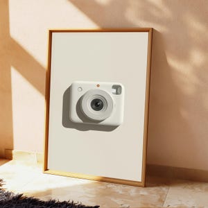 May include: A rectangular wooden frame containing a white print of a white instant camera. The camera features a centered circular lens, a small circular button on the top left, and a rectangular viewfinder window on the top right side.
