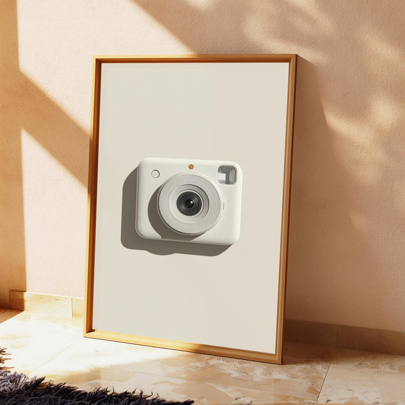 May include: A rectangular wooden frame containing a white print of a white instant camera. The camera features a centered circular lens, a small circular button on the top left, and a rectangular viewfinder window on the top right side.