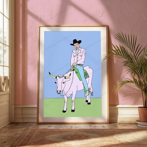 May include: A framed art print featuring a line drawing of a person wearing a black cowboy hat, a light pink jacket, and blue jeans, riding a large light pink cow. The background is a solid light blue sky above a flat green grassy field.