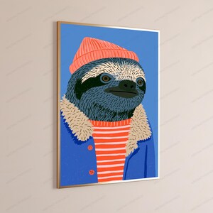 May include: A framed wall art print featuring a stylized illustration of a sloth wearing a coral-colored beanie, a red and white striped shirt, and a blue jacket with a beige faux-fur collar, set against a solid blue background.
