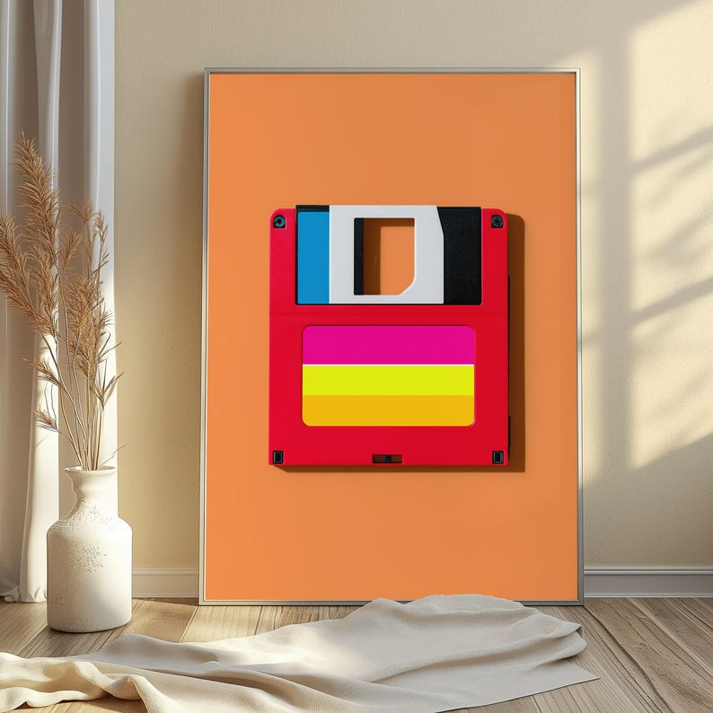 May include: A framed wall art print featuring a red floppy disk graphic centred on a solid orange background. The disk has a blue and black top section with a white square cutout, and a central label area with horizontal pink, yellow, and orange stripes.