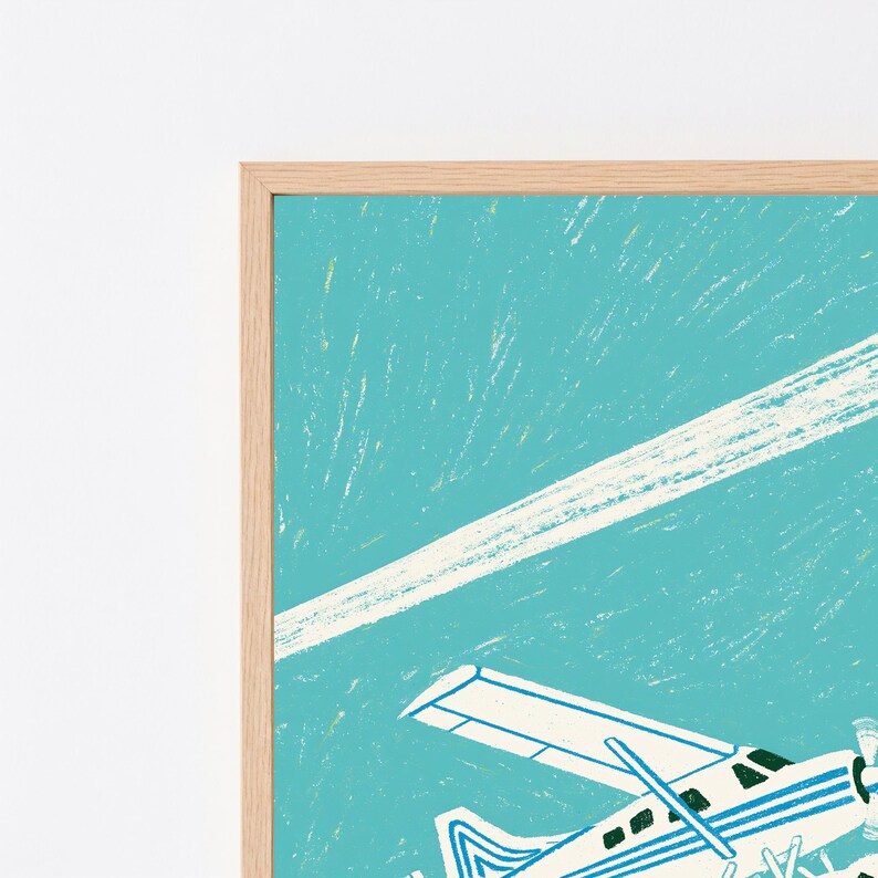 May include: A framed wall art print featuring a white airplane with blue stripes flying against a textured turquoise sky. A long, diagonal white contrail stretches across the upper portion of the print. The frame is made of light-colored wood.