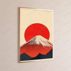 May include: A framed wall art print featuring a stylized mountain with a snow-capped peak against a large, solid red circular sun. The lower portion of the mountain is rendered in deep charcoal tones, while the upper slopes are white and red.