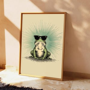May include: A framed wall art print featuring a green frog wearing black sunglasses. The frog is centered against a light beige background with a radiating teal sunburst pattern behind it. The illustration uses bold lines and shading.