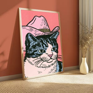May include: A framed wall art print featuring a graphic illustration of a black and white cat wearing a pink cowboy hat. The cat has a white face and chest with black markings around the ears and eyes, set against a solid pink background.