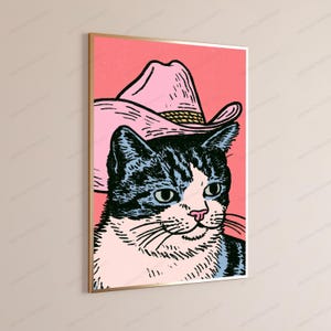 May include: A framed wall art print featuring a graphic illustration of a black and white tabby cat wearing a light pink cowboy hat. The cat is set against a solid coral pink background, rendered in a bold, stylized, and high-contrast artistic style.