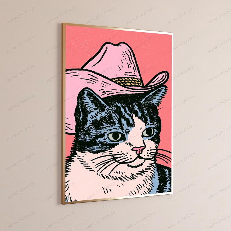 May include: A framed wall art print featuring a graphic illustration of a black and white tabby cat wearing a light pink cowboy hat. The cat is set against a solid coral pink background, rendered in a bold, stylized, and high-contrast artistic style.