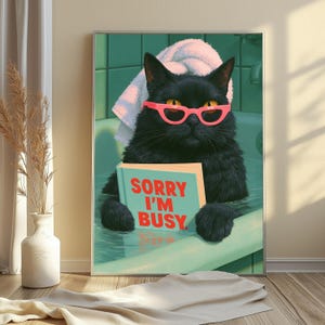 May include: A framed wall art print featuring a black cat with yellow eyes wearing bright pink cat-eye glasses and a white towel on its head. The cat is sitting in a green bathtub holding a light blue book with the red text SORRY I'M BUSY.