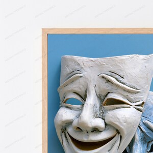 May include: A framed wall art piece featuring a white, textured theatrical comedy mask with a wide, smiling expression. The mask is set against a solid light blue background and enclosed within a light-toned wooden frame with a clean, simple edge.