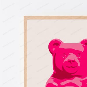 May include: A framed wall art print featuring a vibrant hot pink gummy bear illustration centered on a light beige background. The artwork is enclosed in a light-toned wooden frame, showcasing a clean, graphic design with bold color contrast.