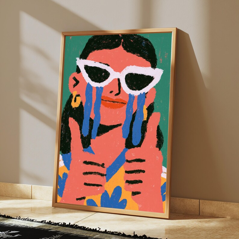 May include: A framed art print featuring a stylized illustration of a person with dark hair wearing white cat-eye sunglasses. The person has blue tear-like shapes streaming from their eyes, a coral-colored face, and is giving two thumbs up.
