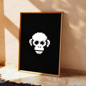 May include: A rectangular framed wall art piece featuring a white pixelated monkey face centered on a solid black background. The frame is made of light-colored wood, and the artwork displays a digital, blocky aesthetic with high contrast.