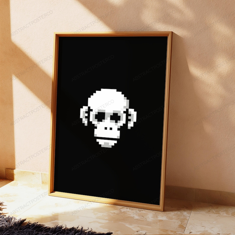 May include: A rectangular framed wall art piece featuring a white pixelated monkey face centered on a solid black background. The frame is made of light-colored wood, and the artwork displays a digital, blocky aesthetic with high contrast.