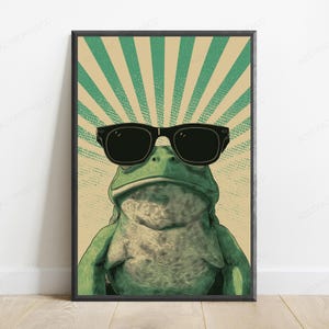 May include: A framed wall art print featuring a green frog wearing black sunglasses. The background consists of a beige and green sunburst pattern with a dotted texture. The frog is centered, facing forward, with a textured skin appearance.