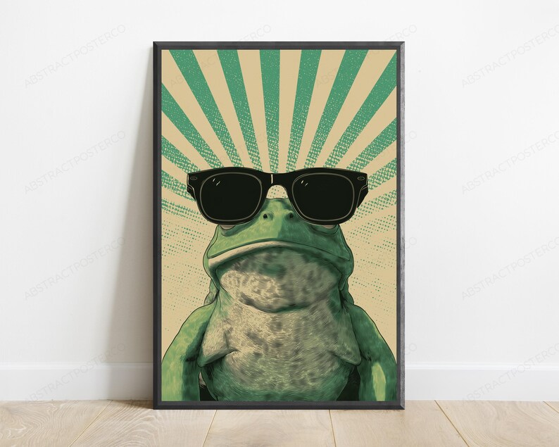 May include: A framed wall art print featuring a green frog wearing black sunglasses. The background consists of a beige and green sunburst pattern with a dotted texture. The frog is centered, facing forward, with a textured skin appearance.