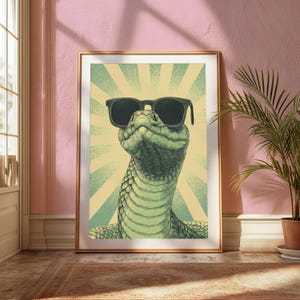May include: A framed wall art print featuring a detailed illustration of a green snake wearing black sunglasses. The snake is positioned centrally against a background of light yellow and green radiating sunburst lines in a vintage graphic style.