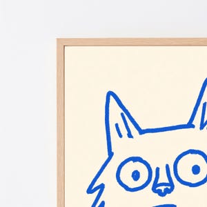 May include: A framed wall art print featuring a bold blue line drawing of a cat face with large circular eyes and pointed ears on a cream-colored background. The artwork is encased in a light-toned wooden frame with a clean, simple border design.