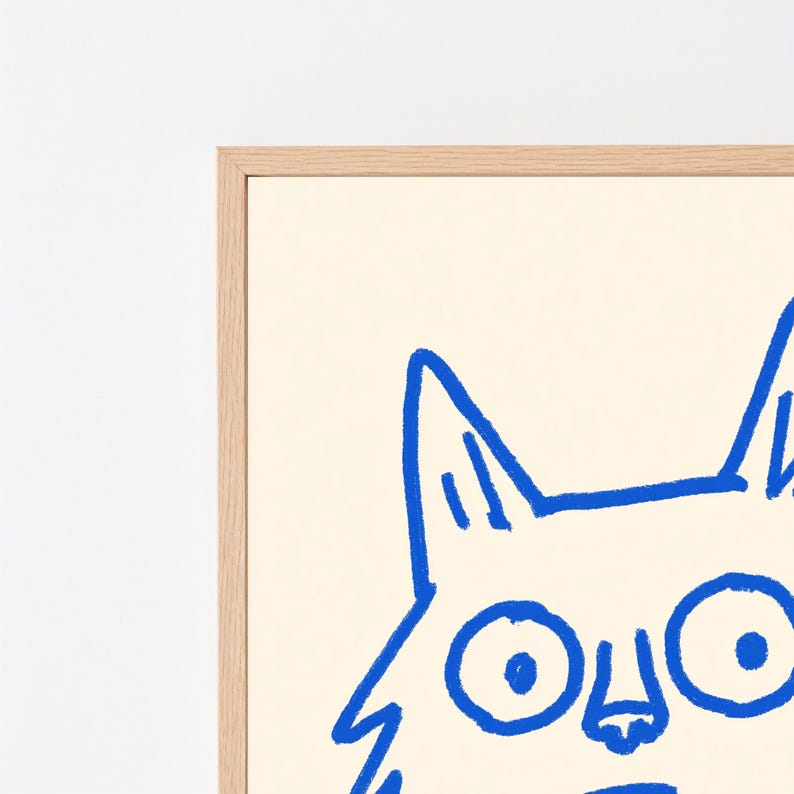 May include: A framed wall art print featuring a bold blue line drawing of a cat face with large circular eyes and pointed ears on a cream-colored background. The artwork is encased in a light-toned wooden frame with a clean, simple border design.