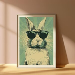 May include: A framed wall art print featuring a detailed illustration of a rabbit wearing black sunglasses. The rabbit is centered against a background of radiating light green and cream-colored sunburst lines, creating a bold graphic effect.