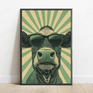 May include: A framed wall art print featuring a dark-colored cow wearing black sunglasses. The cow is centered against a background of alternating light green and cream-colored sunburst rays. The artwork has a detailed, textured illustration style.