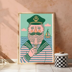 K&ouml;nnte beinhalten: Ein gerahmter Kunstdruck zeigt einen b&auml;rtigen Seemann mit einer dunkelgr&uuml;nen Kapit&auml;nsm&uuml;tze mit goldenem Ankeremblem und einem rosa-blau gestreiften Hemd. Der Seemann isst ein Eis vor einem hellgr&uuml;nen Hintergrund mit einem kleinen Segelboot.