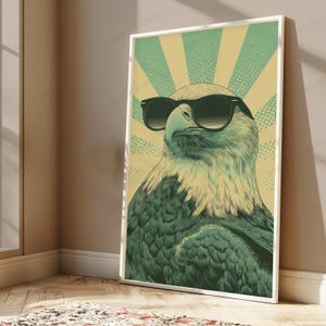 May include: A framed vertical wall art print featuring a stylized illustration of a bald eagle wearing black sunglasses. The eagle is rendered in shades of dark green and cream, set against a background of radiating cream and green sunburst rays.