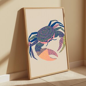 May include: A framed wall art print featuring a stylized crab illustration on a light beige background. The crab is rendered with a mix of blue, pink, and yellow patterns, including dots and stripes, contained within a simple light wood frame.