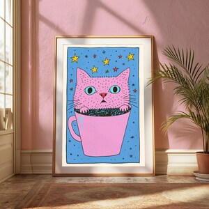 Pu&ograve; includere: Una stampa artistica incorniciata raffigurante un gatto rosa con grandi occhi azzurri seduto all'interno di una tazza rosa. Lo sfondo &egrave; di un blu vivace, costellato di stelle gialle e rosse. L'illustrazione presenta contorni neri e un tratto.