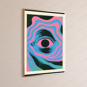 May include: A rectangular wall art print in a gold frame featuring a central black pupil surrounded by a pink iris. The eye is set against a background of wavy, concentric lines in alternating shades of bright pink, light blue, and dark teal.