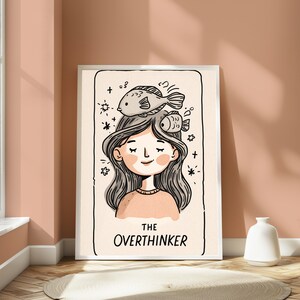 K&ouml;nnte beinhalten: Ein rechteckiger Wandkunstdruck mit einer schwarz-wei&szlig;en Strichzeichnung einer Person mit langem Haar und zwei Fischen auf dem Kopf. Der Hintergrund ist in einem sanften Cremeton gehalten und der Text THE OVERTHINKER steht unten.