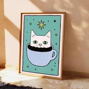 May include: A framed rectangular wall art print featuring a white cat with green eyes sitting inside a light blue teacup filled with black liquid. The background is teal with a pale pink border, decorated with small yellow stars and black cross symbols.