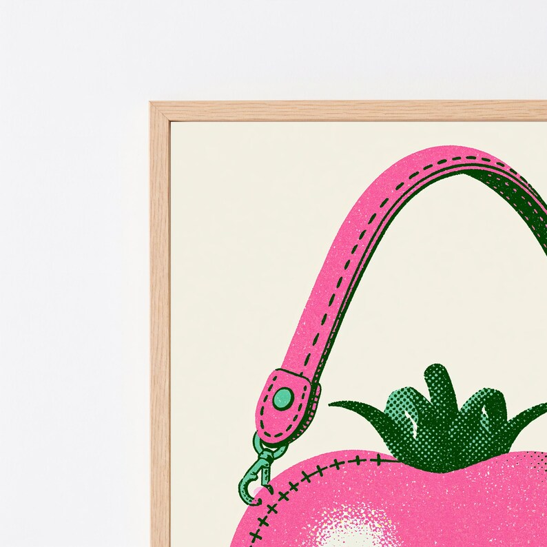 May include: A framed wall art print featuring a stylized pink tomato-shaped handbag with a bright pink strap, a green stem, and dark green stitching details. The illustration uses a dotted halftone texture and is set within a light wood frame.