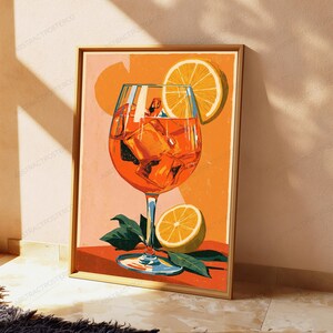 May include: A framed art print featuring a vibrant orange cocktail in a stemmed glass filled with ice cubes. A bright yellow lemon slice garnishes the glass, while another lemon slice and green leaves rest at the base on an orange surface.